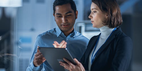 Zwei Personen in modernem Büro betrachten ein Tablet; eine Person zeigt darauf.