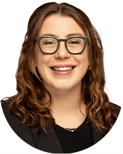 A woman with shoulder-length wavy hair, wearing glasses, a black top, and a dark blazer smiles at the camera.