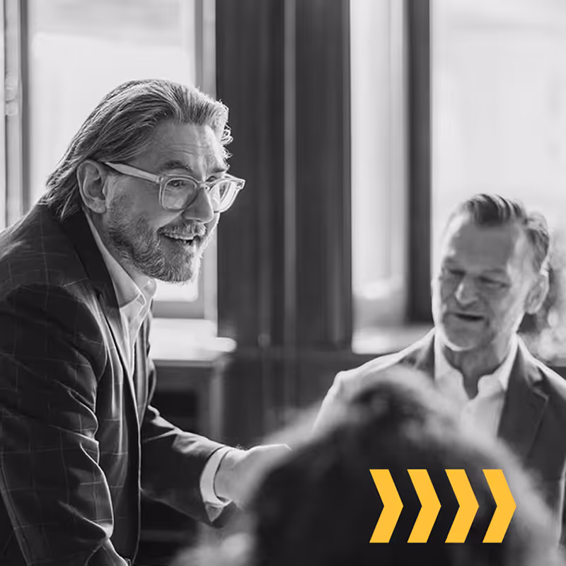 Two men in suits engaged in conversation in a bright office with large windows, with yellow arrows overlay.