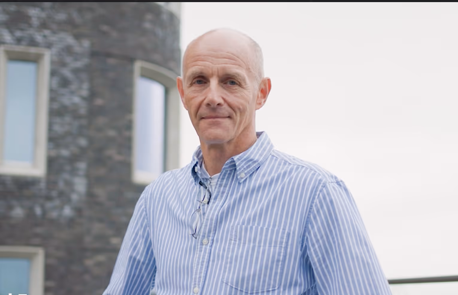 Middle-aged man wearing a blue and white striped shirt standing outdoors in front of a stone building.
