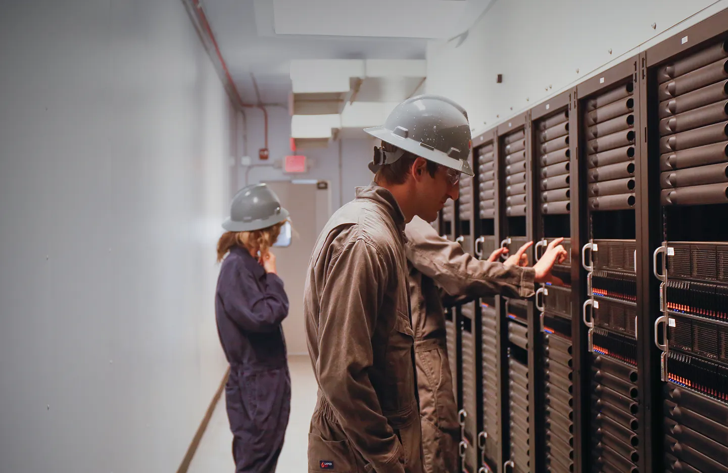 Three workers wearing hard hats and uniforms inspecting server racks in a data center hallway.