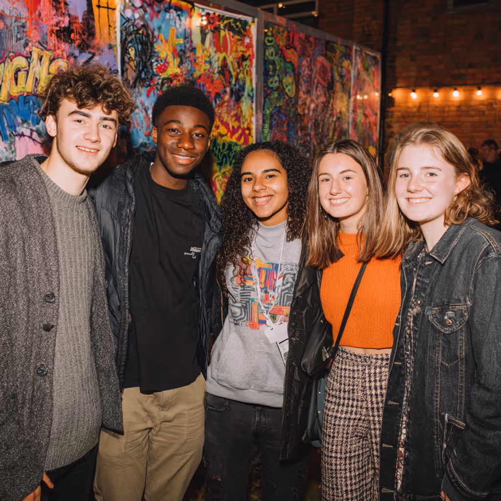 Group of five young people smiling and posing in front of a colorful graffiti wall at an event.