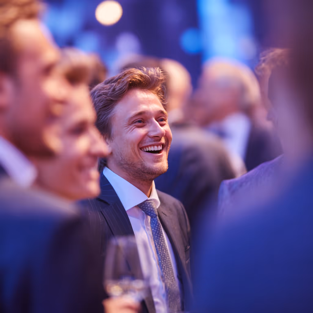 Smiling businessman in suit and tie enjoying a lively networking event with blurred people and warm lighting in the background.
