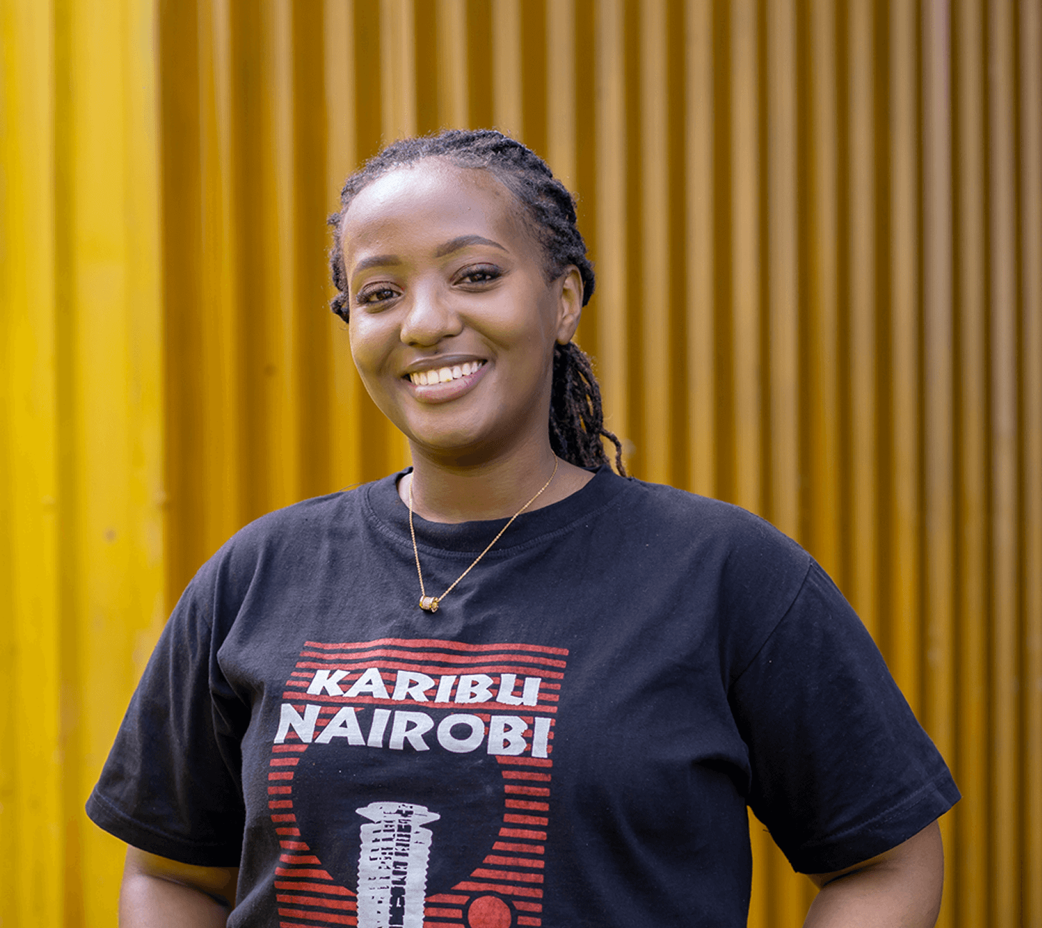 Smiling woman with braided hair wearing a black T-shirt that says 'Karibu Nairobi' standing in front of a yellow corrugated wall.