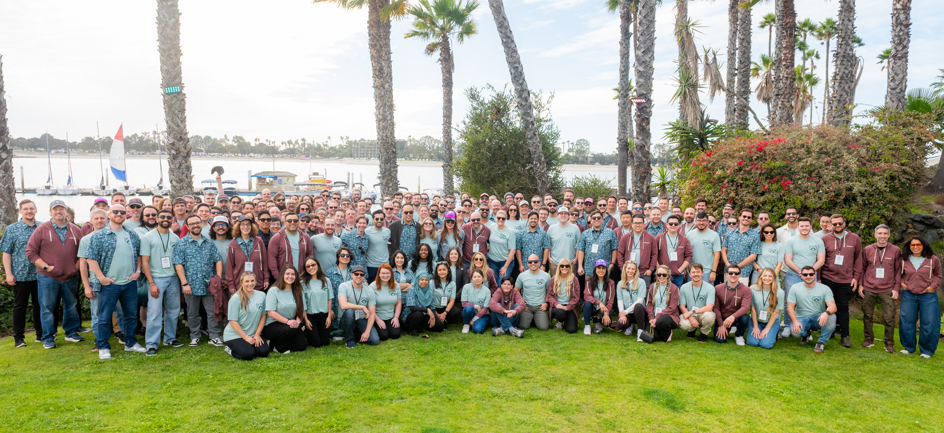 Large group of people posing outdoors on green grass with palm trees and boats on water in the background.