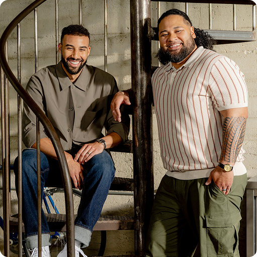 Two Shopify merchants smiling on a spiral staircase. One wears a gray shirt and jeans, while the other is in a striped polo and green pants.