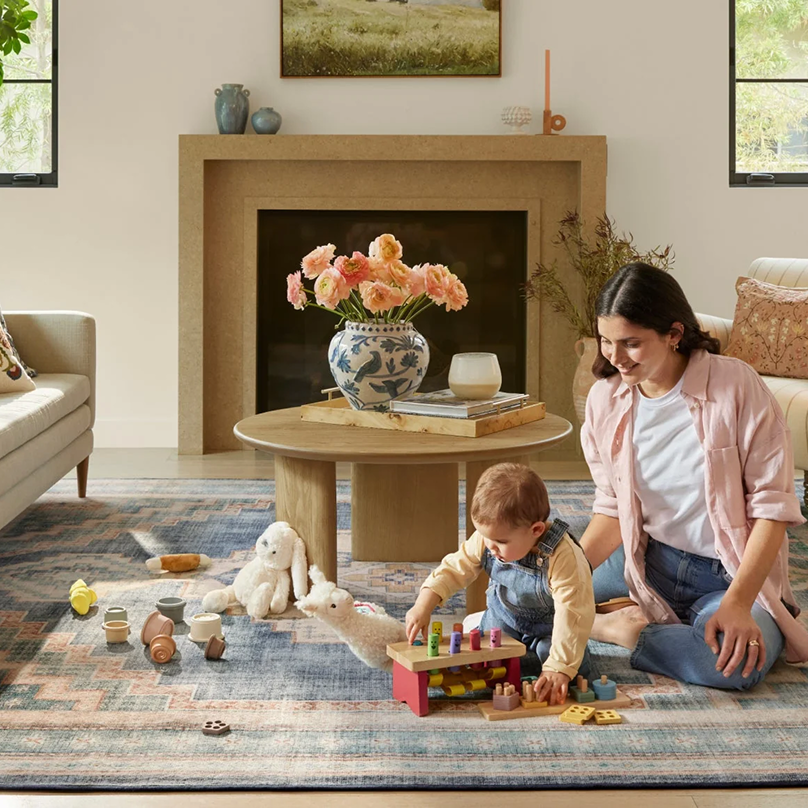 Frau und Baby spielen mit Holzspielzeug auf einem Wohnzimmerteppich.
