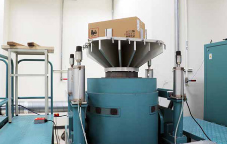 Thiết bị kiểm tra độ rung công nghiệp lớn với hộp các tông ở trên cùng, xung quanh là máy móc màu xanh và lan can an toàn.