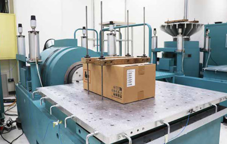 Thiết lập thiết bị công nghiệp trong môi trường phòng thí nghiệm với nhiều loại máy móc, bảng điện và cáp được kết nối với một hộp các tông lớn trên giá đỡ.