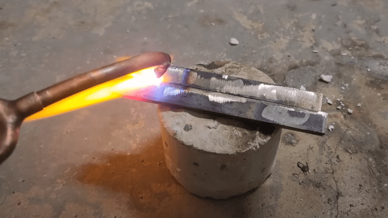 Two pieces of metal are sitting next to each other on a brick, with one edge touching. The copper end of a torch is directing a flame against the metal, and the metal is glowing brightly around the point where the flame contacts it.