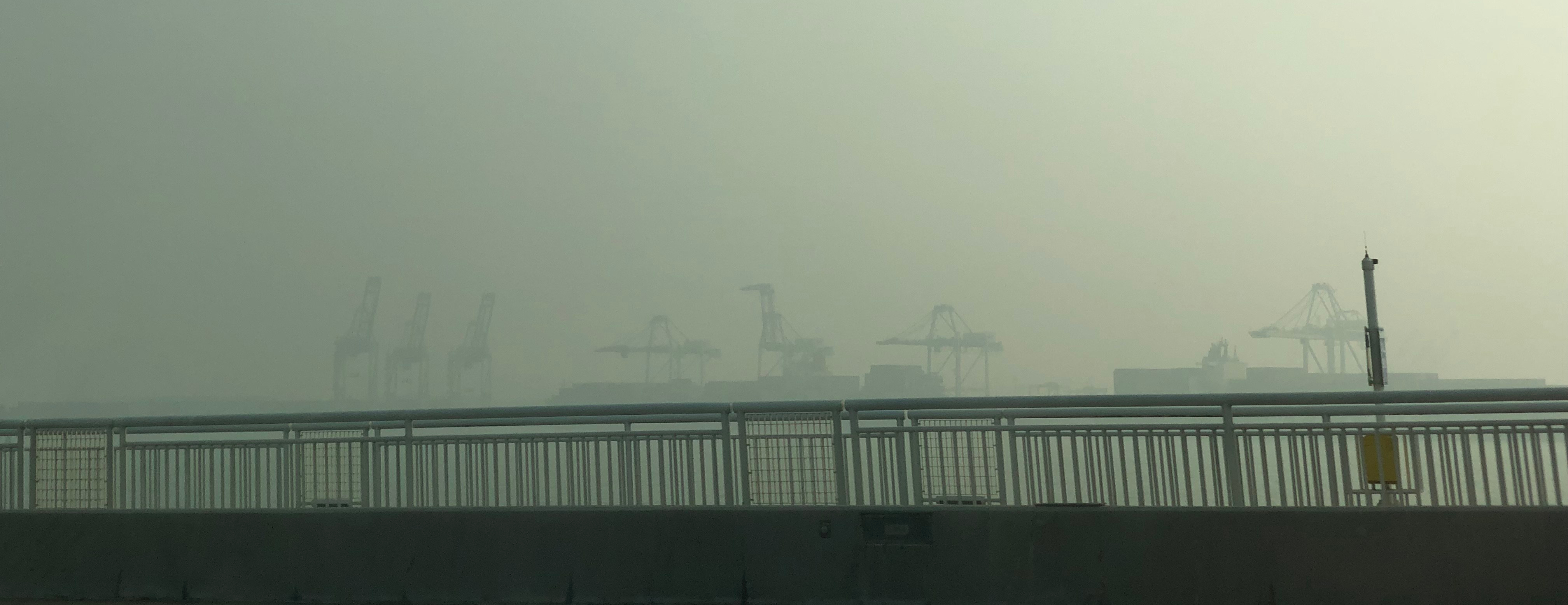 shipping container cranes are hidden by smoke in the distance beyond the bridge's railing
