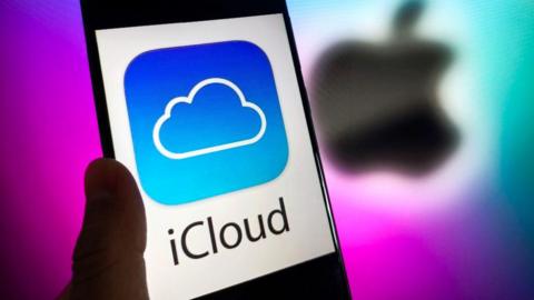A phone with the words iCloud in black letter, and the iCloud blue and white logo of a cloud on it, against a white background. A blurred Apple logo in black is behind it against a purple background.
