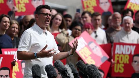 Burnham, wearing a white polo t-shirt, speaks into microphones while many people stand behind him holding red signs that say 'Andy for us'