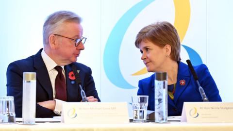 Michael Gove and Nicola Sturgeon sitting next to each other at a table. Gove has his arms closed, Sturgeon is leaning towards him. They are looking at each other directly in the eye, stony-faced.