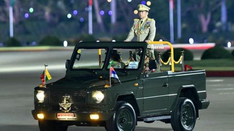 Myanmar's junta chief military Min Aung Hlaing arrives to deliver a speech during a ceremony to mark the country's Armed Forces Day in Naypyidaw in 2024. He is wearing a military outfit and standing in an open top green car.