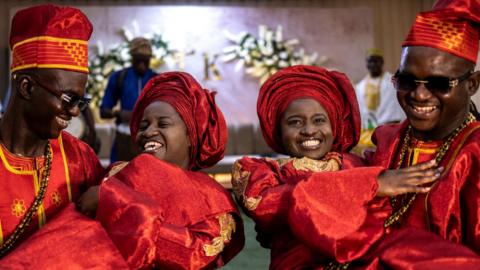 Twin brothers Kehinde and Taiwo Oguntoye carry Kehinde and Taiwo Adediran. Both couples wear red traditional formalwear.