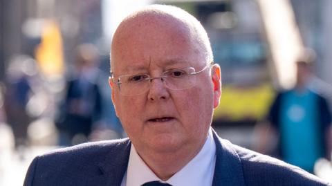 Peter Murrell, who is bald with glasses, is pictured on a busy street looking deep in thought