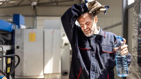 A man in overalls wiping his forehead and holding a plastic water bottle