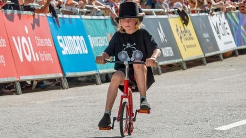 A young girl in a black bowler-style hat and shorts and t shirt rides towards the camera on a small, red penny farthing in London as crowds smile in the background behind barriers. The weather is sunny.