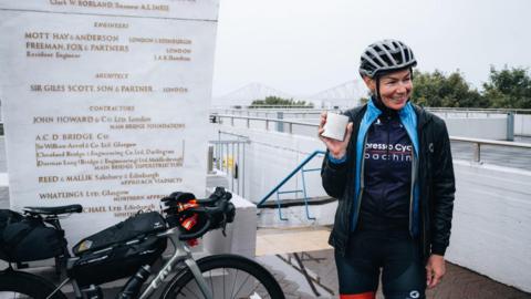 Cyclist Christina MacKenzie to the right of screen in black cycling attire, holding a her winning silver hip flask filled with irn-bru. Her grey Liv bike is to the left of screen. Photo taken at South Queensferry Memorial.