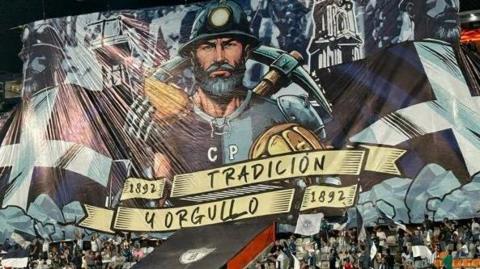 A large flag in a Mexican football stadium showing a Cornish miner holding a pasty and a pickaxe with two Cornish St Pirans flags either side of him. The flag shows the year 1892, when Pachuca were founded, as well as text that says 'tradicion y orgullo' - meaning tradition and pride in English.