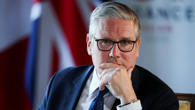 Keir Starmer sits with his hand covering his mouth. He is wearing a suit with a union jack in the blurred background.