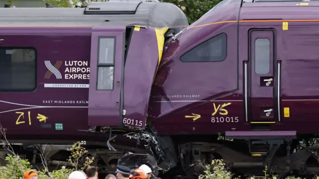 An image of the crash. Two trains together on the track, the front of one is partially covered by the debris of the other.