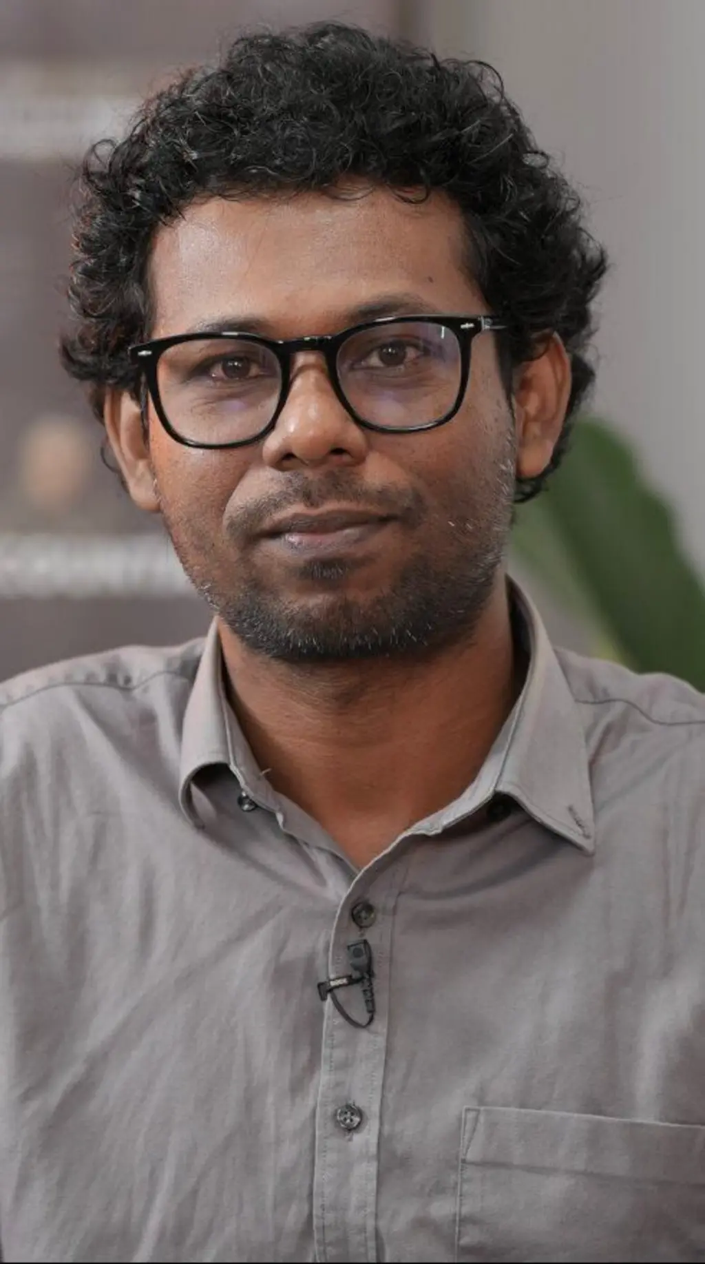 ක්රේල් කොණ්ඩයක් සහිත, කණ්නාඩි පැළඳ සිටින අයෙකු අළු පාට කමිසයක් ඇඳ සිටී.