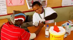 A woman in a township is testing another person, holding their hand. There are Aids posters in the background and a sharps container on the table.