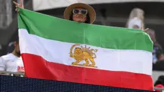 2026 йил 15 июнь куни Инглвуд шаҳридаги Лос-Анжелес стадионида Эрон ва Янги Зеландия ўртасида бўлиб ўтган Жаҳон чемпионати–2026нинг G гуруҳи баҳси олдидан бир эронлик мухлис инқилобдан олдинги Эрон байроғини намойиш этмоқда.