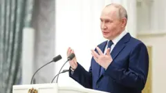 Оқ кўйлак, кўк нуқтали галстук ва кўк костюм кийган президент Путин икки микрофон ўрнатилган оқ минбар ортида нутқ сўзлар экан, қўл ҳаракатлари билан фикрини ифода этмоқда. Суратнинг чап томонида оч яшил рангли нақшли парда кўриниб турибди.