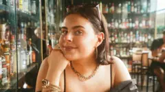 A young woman in a bar with lots of bottles of drink in cabinets behind her. You can see the top half of her body. She is wearing a black top and resting her chin on her hand. She has sunglasses pinning back her hair and has golden earings, necklace and bracelet.