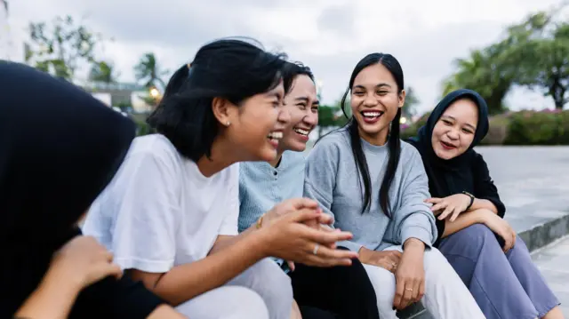 A group of five female teenagers sit on a pavement together smiling and laughing