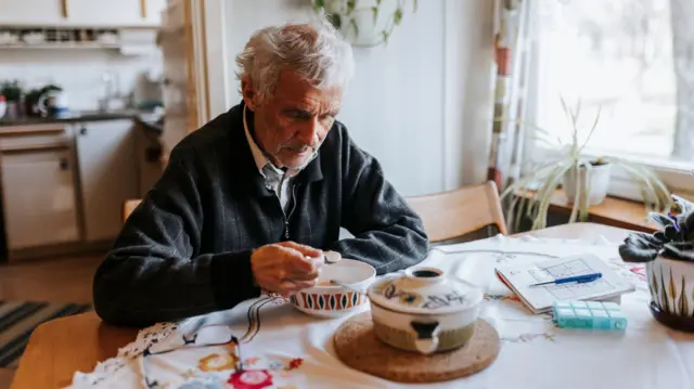 An older man with grey hair sits at a table in his home eating cereal from a bowl.