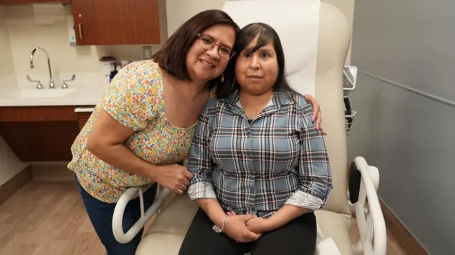 Jessica Lopez sitting in a hospital chair wearing a black trouser and a shirt getting a warm embrace from her mum