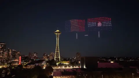 The Seattle skyline at night with drones lights showing flags and 1-1
