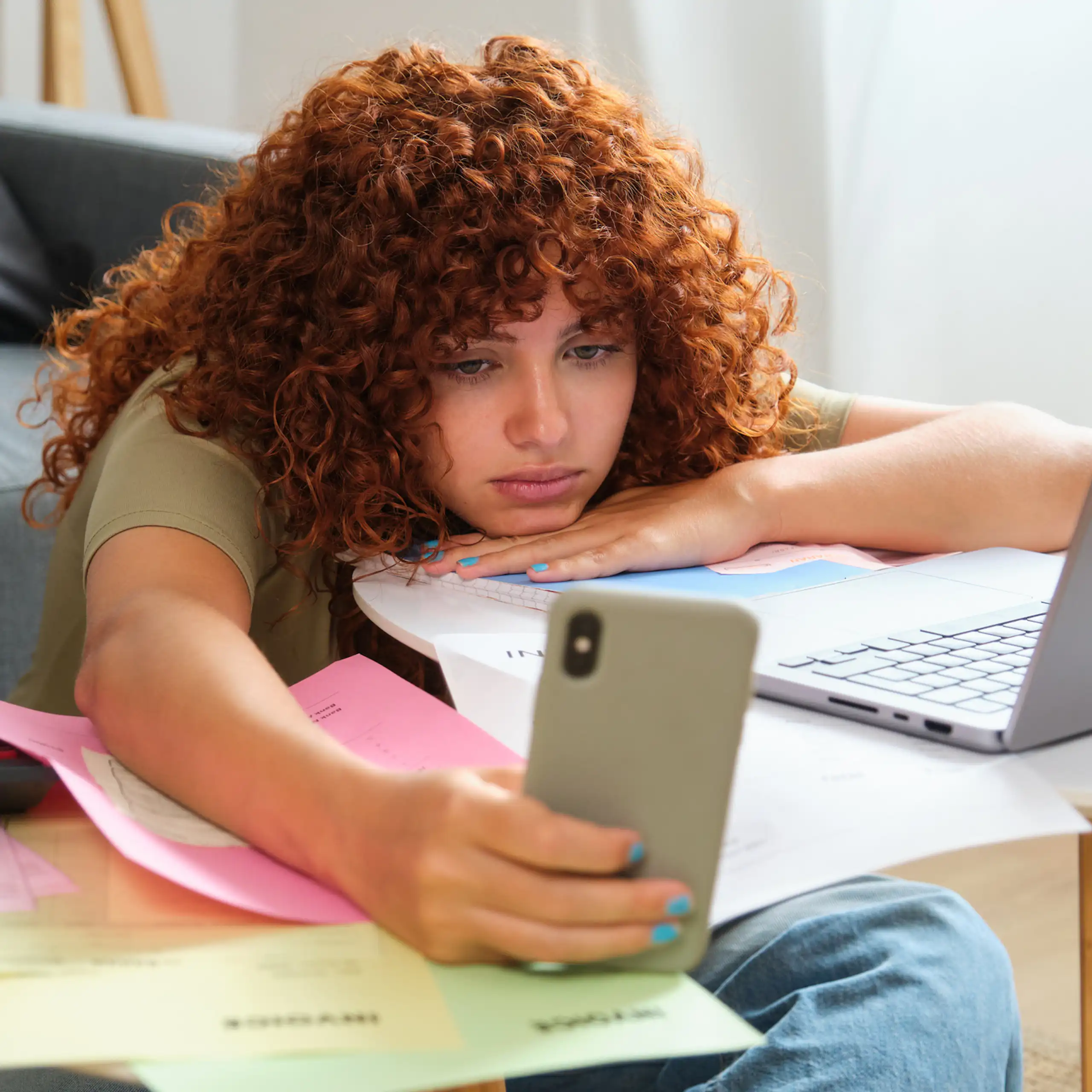 Young woman looks at phone while sitting at a table with a laptop and paperwork.