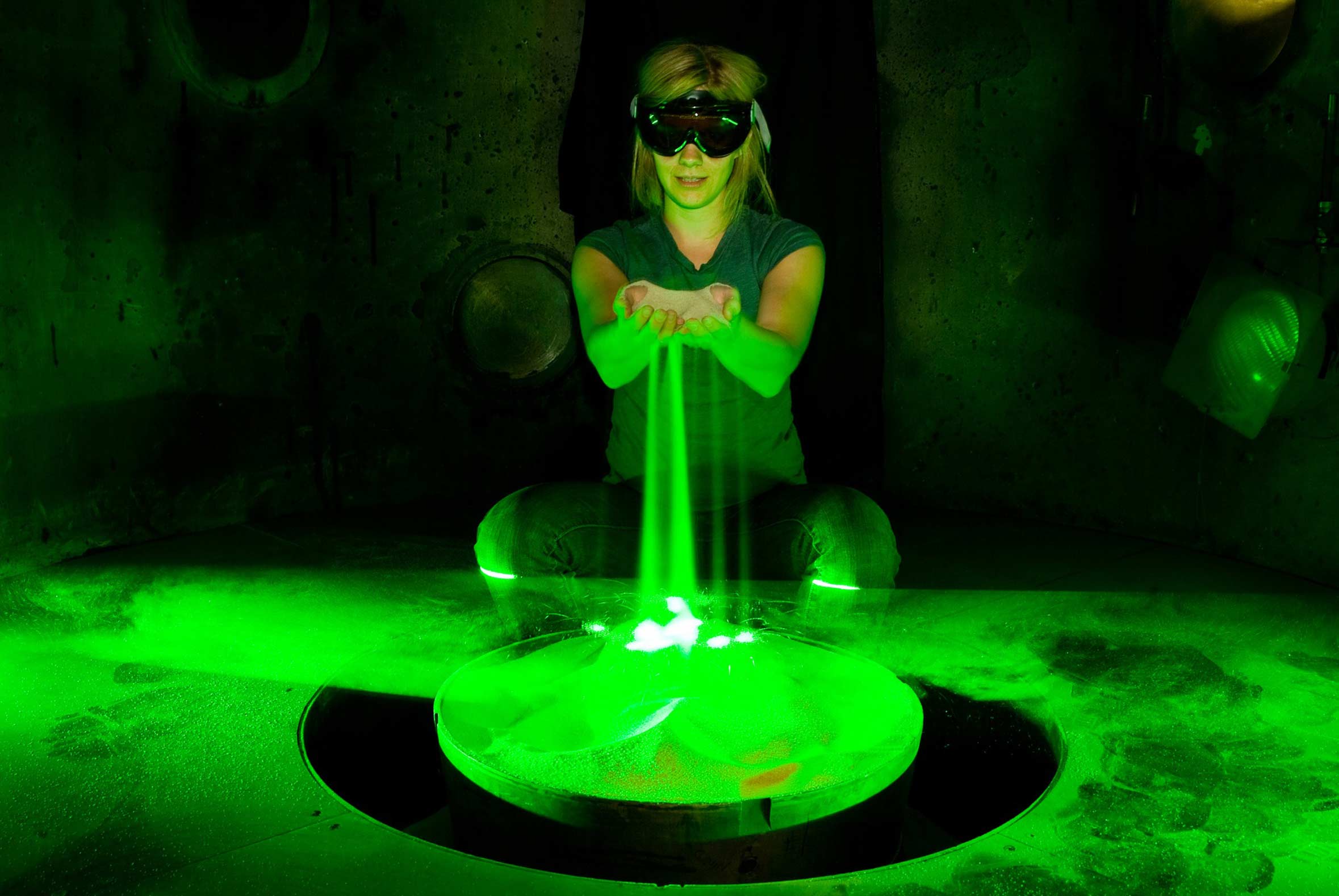 Sand glowing green from lasers slipping between hands of woman in dark room wearing goggles.